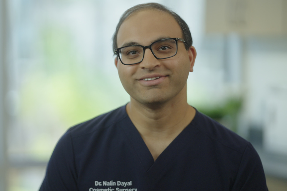 Smiling person wearing glasses and a navy scrub top with text, in a well-lit room with blurred background.