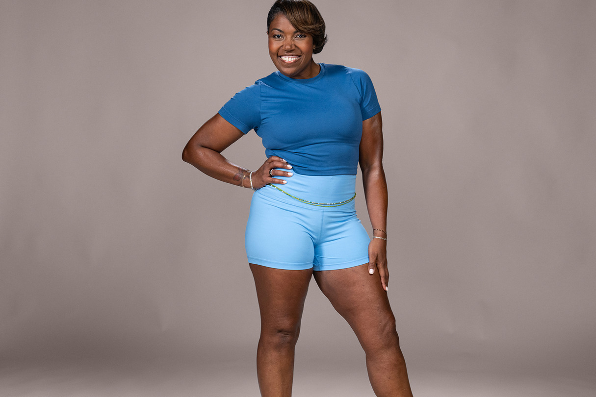A smiling woman in a blue workout outfit poses confidently against a neutral background.