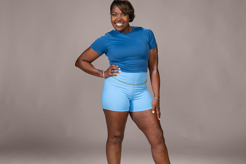 A smiling woman in a blue workout outfit poses confidently against a neutral background.