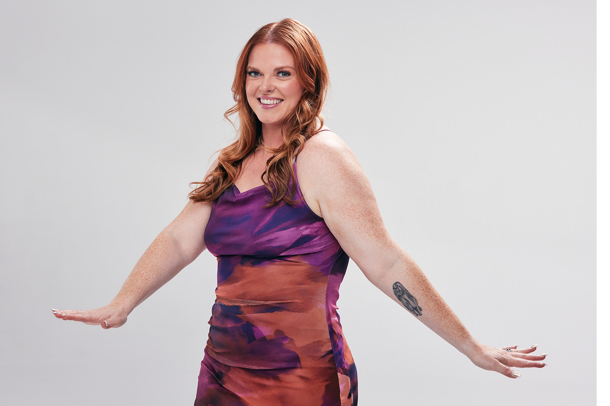 Person wearing a colorful dress poses against a neutral background, showcasing a forearm tattoo while smiling.