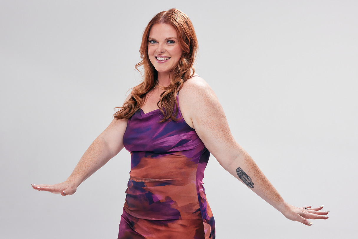 Person wearing a colorful dress poses against a neutral background, showcasing a forearm tattoo while smiling.