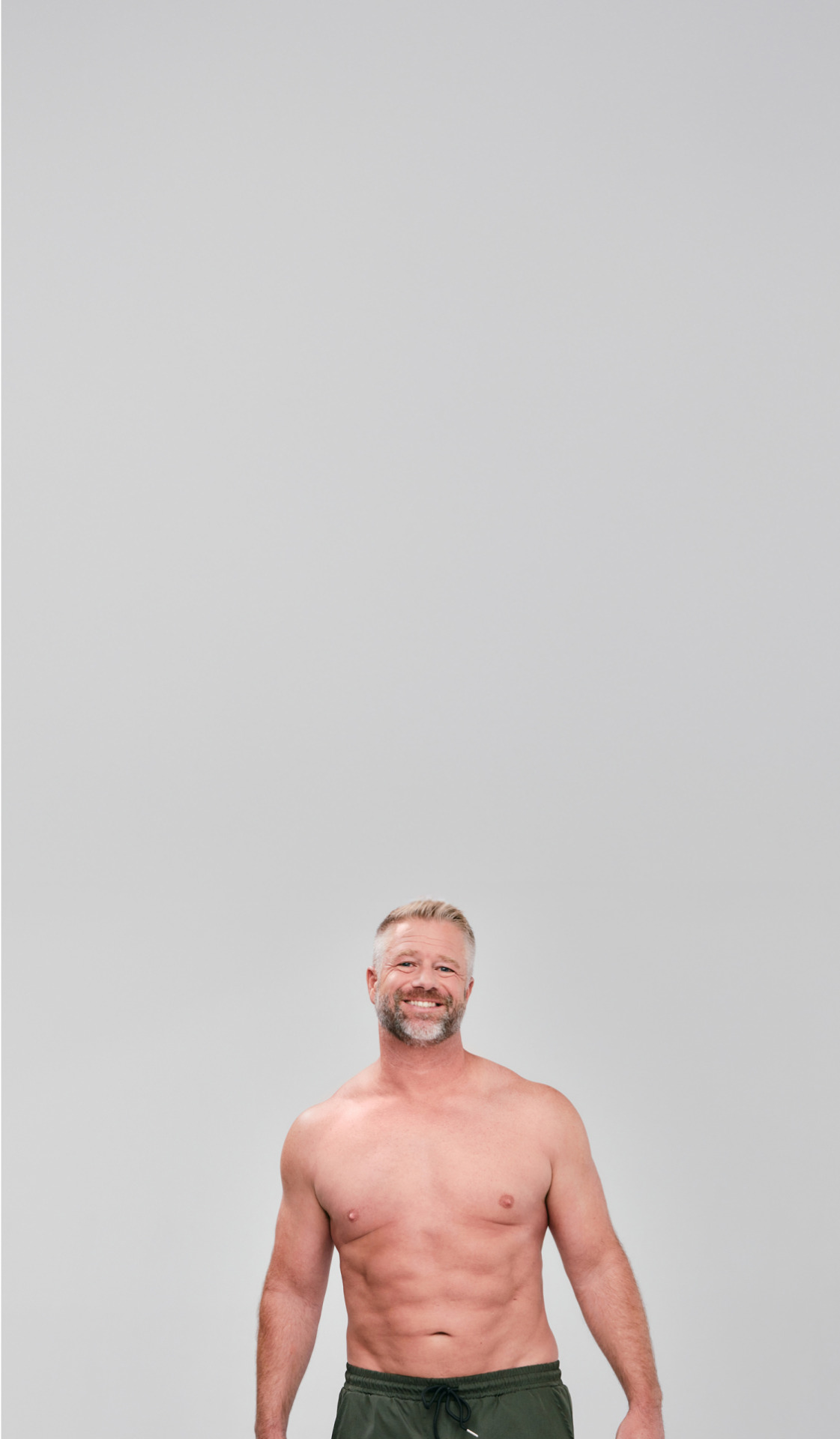 Smiling man with short hair, wearing green shorts, standing against a plain gray background.