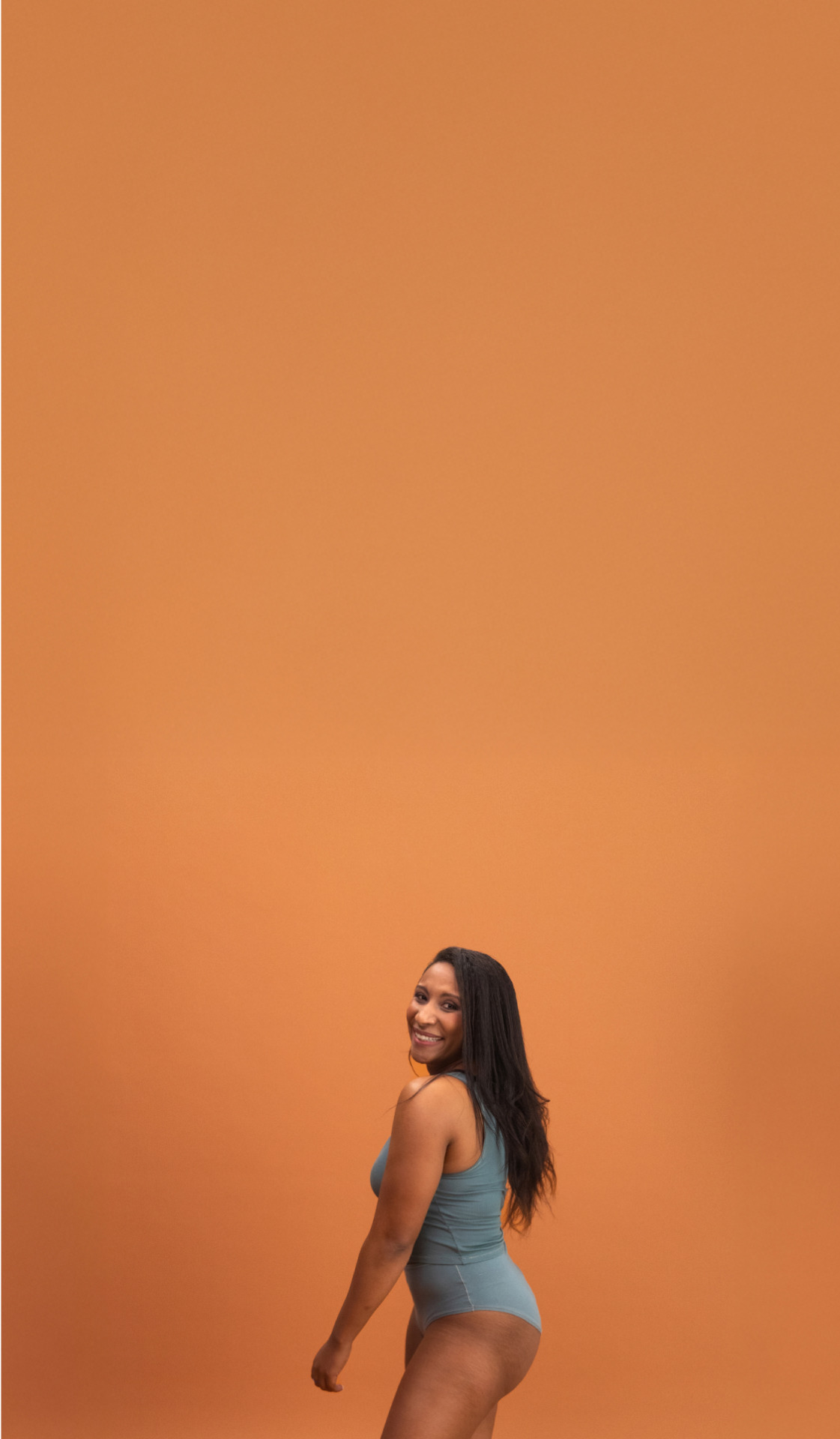 Woman in a teal swimsuit standing against an orange background, smiling and looking over her shoulder.