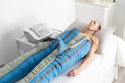 Person undergoing pressotherapy treatment, lying on a therapy table with a massage suit, next to a medical device.