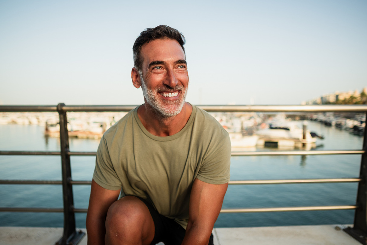 Smiling man sitting on a dock with boats in the background on a sunny day.