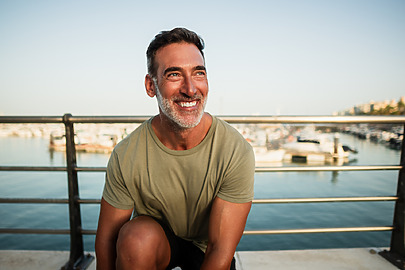 Smiling man sitting on a dock with boats in the background on a sunny day.