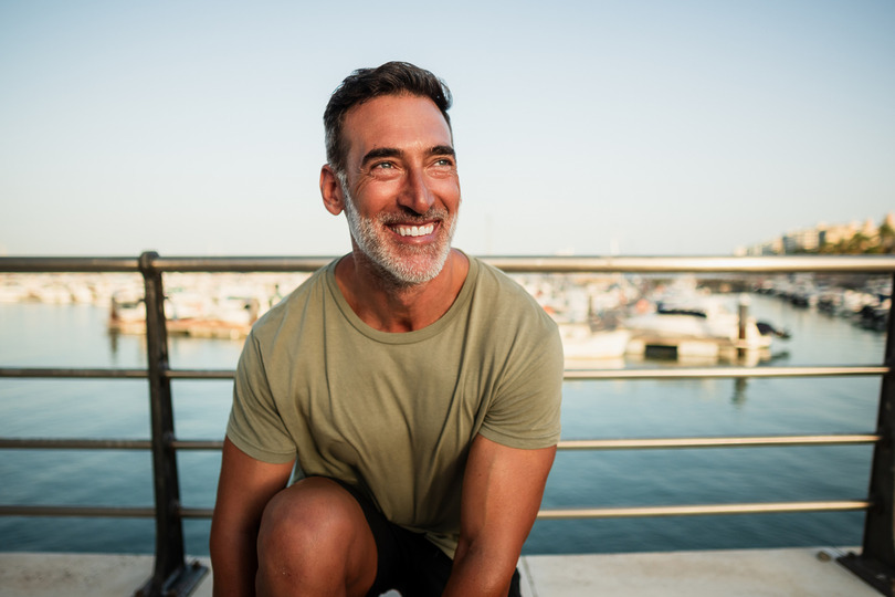 Smiling man sitting on a dock with boats in the background on a sunny day.