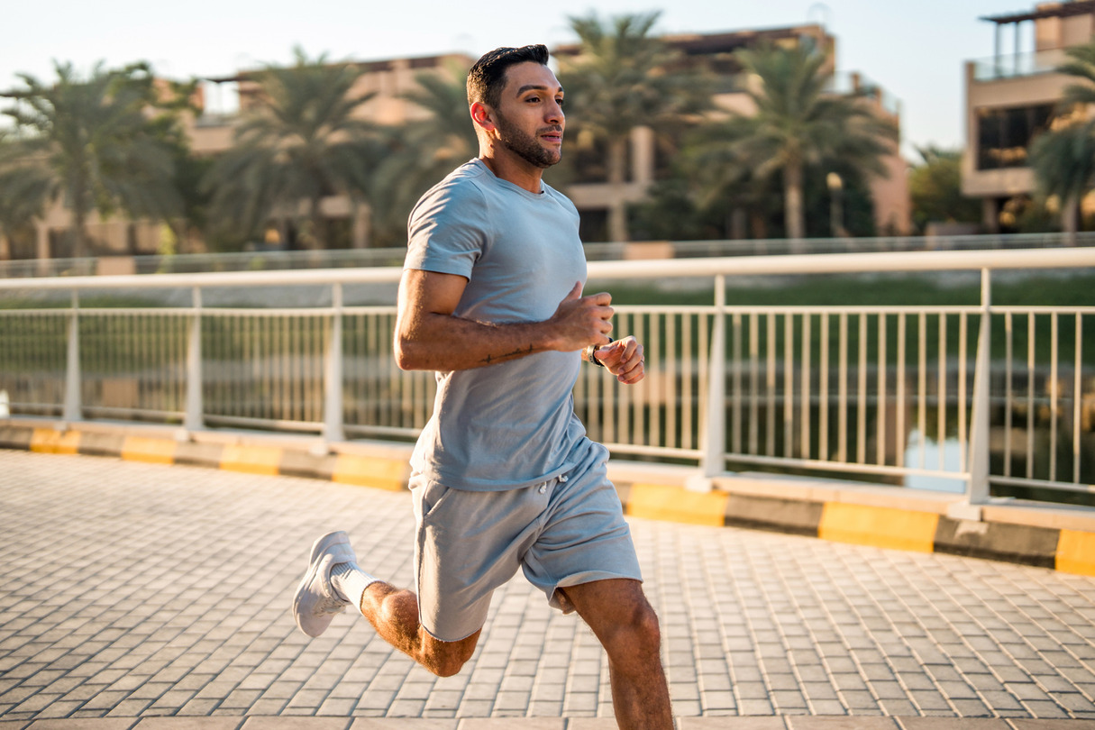 Man jogging on a sunny day, wearing a light gray outfit, with palm trees and buildings in the background.
