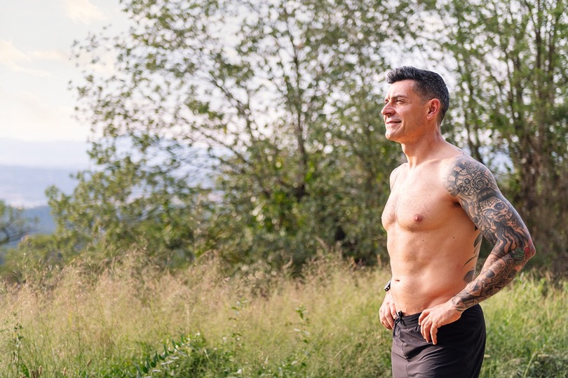 Fit man with tattoos stands outdoors, shirtless, hands on hips, surrounded by greenery, looking content in nature.