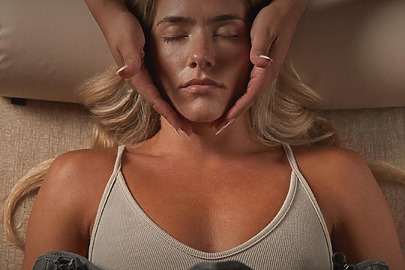 Woman receiving a facial massage while lying down, eyes closed, and wearing a light-colored tank top.