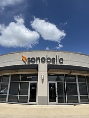 Exterior view of a Sono Bello clinic with logo sign, glass windows, and a sunny blue sky with clouds.