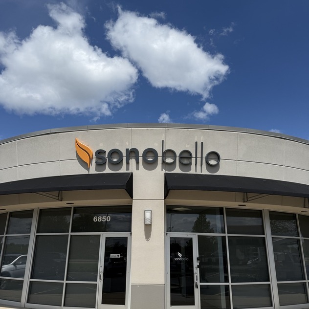 Exterior view of a Sono Bello clinic with logo sign, glass windows, and a sunny blue sky with clouds.