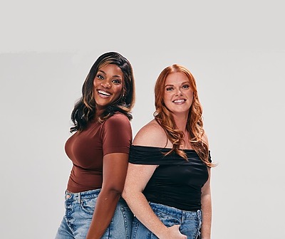 Two women smiling, standing back to back, wearing casual tops and denim jeans against a plain background.